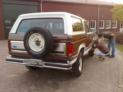 1984 Ford Bronco - Brown and white Ford Bronco from the rear, with two men inspecting the front wheel area.