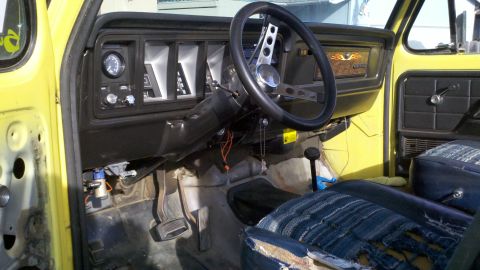 1978 Ford Bronco - The interior of a yellow vehicle shows a dashboard with gauges, an aftermarket steering wheel, and worn seats.