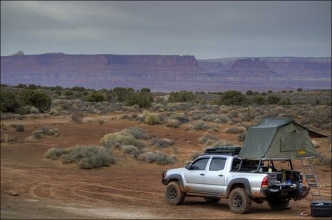 1990 Chevrolet Suburban - A silver pickup truck equipped with a rooftop tent is parked in a dry, desert environment with mesas in the background.