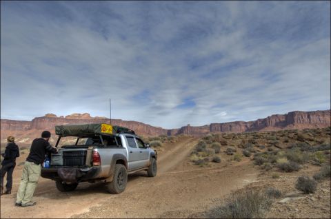 1990 Chevrolet Suburban - A silver pickup truck equipped with a rooftop tent is parked on a dirt road, with people loading gear, set against a backdrop of desert can…
