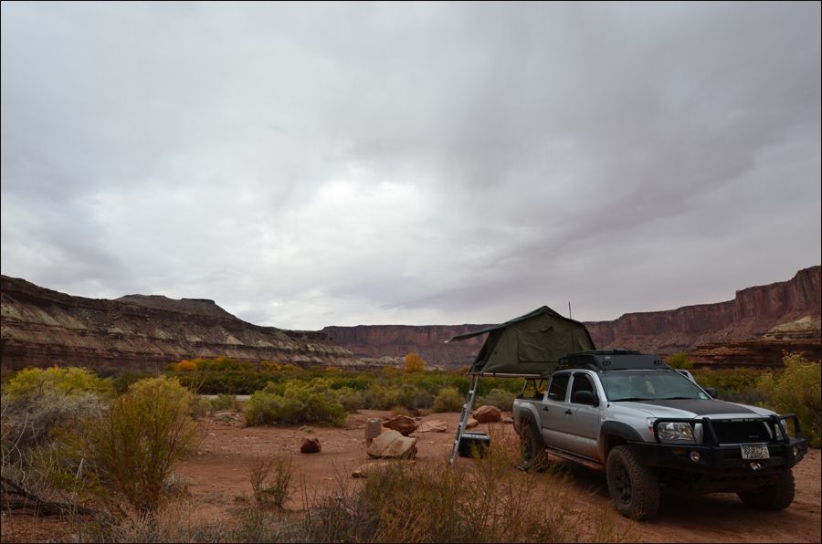 Silver pickup truck with a rooftop tent set up, parked in a desert area with rocky cliffs in the background