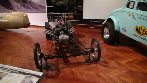 1993 Ford Taurus - A dragster and a vintage race car are displayed indoors on a polished wooden floor.