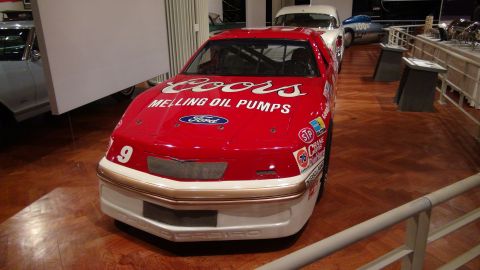 1993 Ford Taurus - A red race car with sponsor decals is displayed indoors, with other vehicles in the background.