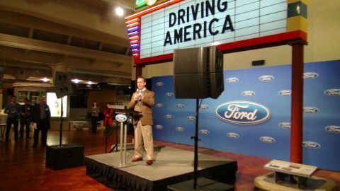 1993 Ford Taurus - A speaker addresses an audience at an event Ford branding and a "DRIVING AMERICA" display.
