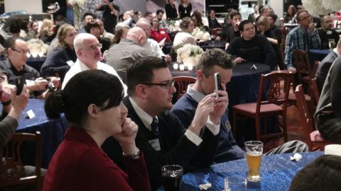 1993 Ford Taurus - Attendees at an indoor event are seated at tables, with some individuals engaged with their mobile devices.