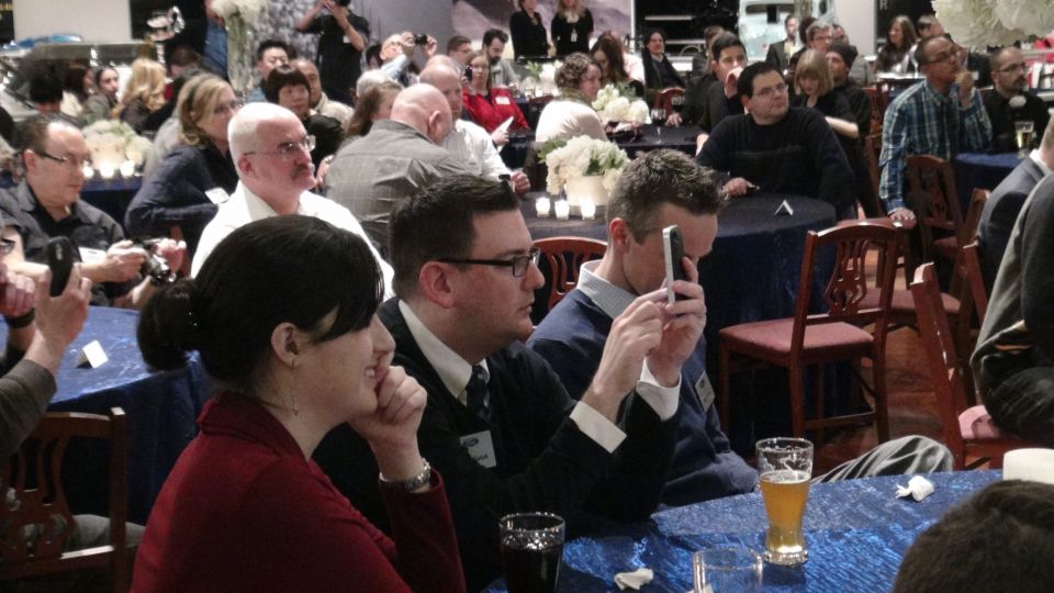 People seated at tables, some looking at phones, at an indoor event