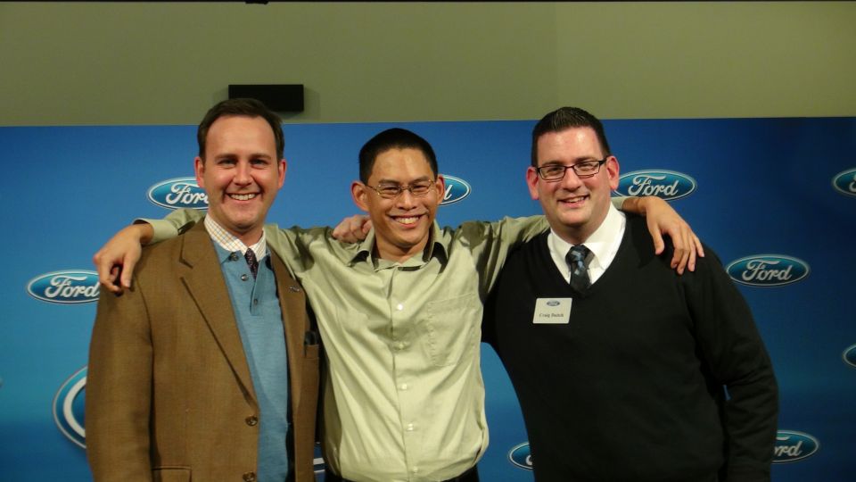 Three men stand with arms around each other in front of a blue backdrop with Ford logos. One man wears a brown jacket, another a light green shirt, and the third a black sweater