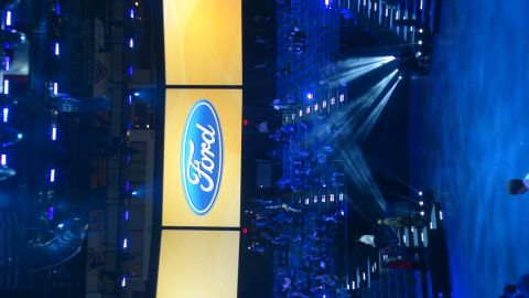 1993 Ford Taurus - The Ford logo is prominently displayed on a screen during an auto show event, with dramatic lighting and an audience visible.