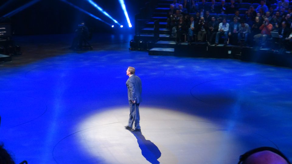 Man in a suit stands on a brightly lit blue stage with an audience in the background
