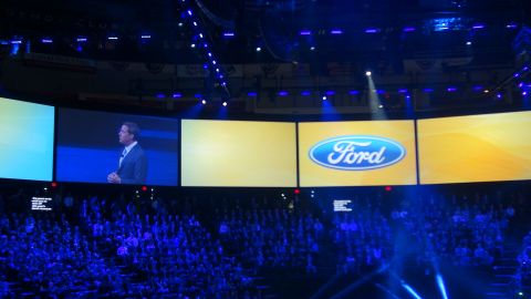 1993 Ford Taurus - A Ford logo is prominently displayed on a screen at an automotive event, with a speaker presenting to a large audience.