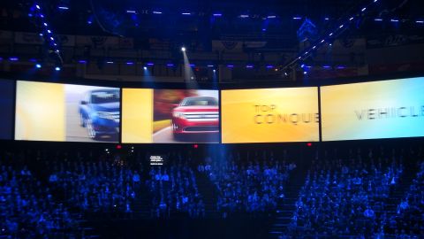 1993 Ford Taurus - A large screen displays multiple vehicles and text during an event attended by a large audience.