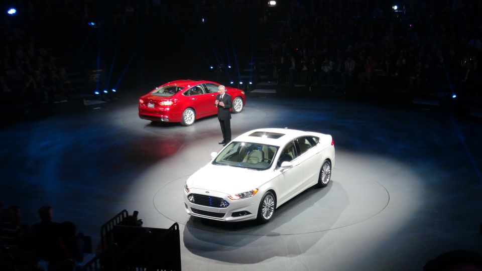 White Ford sedan with a sunroof on a stage, front 3/4 view. A red Ford sedan is behind it