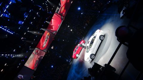1993 Ford Taurus - A white sedan is presented on a stage, with multiple red car images displayed on screens behind it.