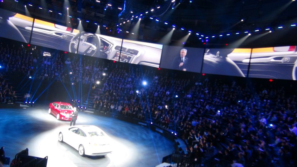 Two cars on stage at an auto show presentation with a large screen showing car interior and a speaker