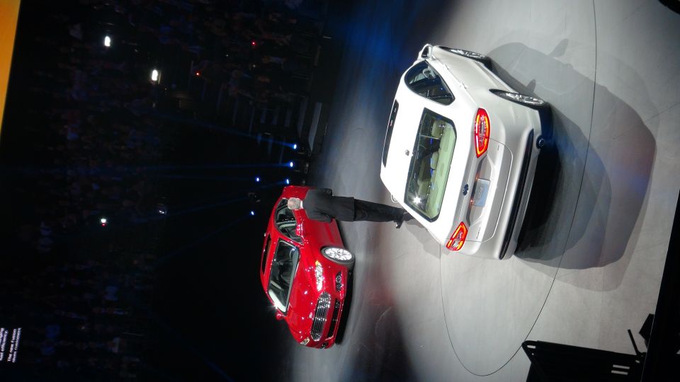 Man in suit next to red car, white car behind him on a stage with audience in background