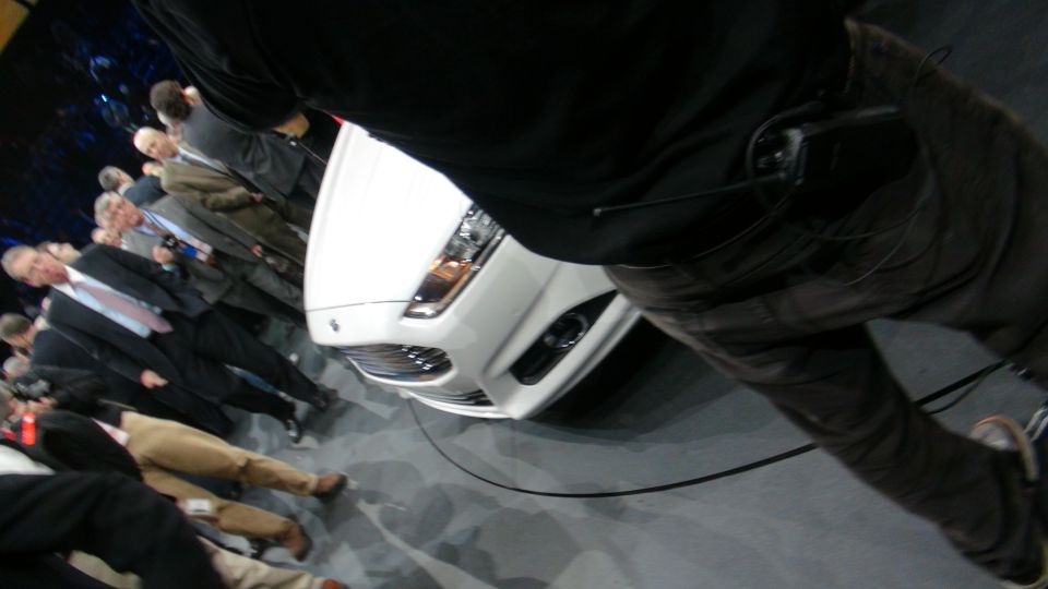 White car front grille and headlights at an auto show, with people in suits around it
