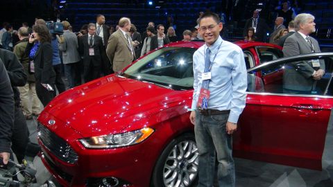 1993 Ford Taurus - A man poses next to a red Ford sedan at an event with other people in the background.