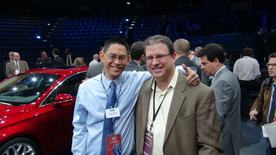 Red car side view with a man in a blue shirt and tie standing next to it