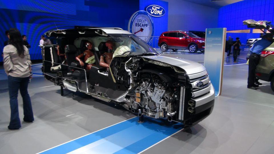 Cutaway Ford Explorer showing interior and engine components, with crash test dummies inside. Other Ford vehicles are visible in the background