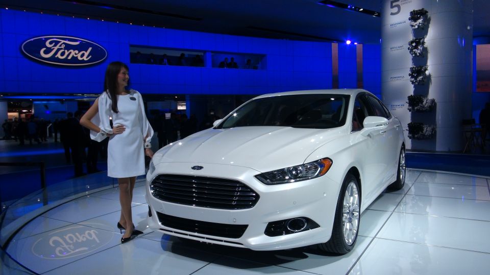 White Ford sedan on display with a woman in a white dress. Ford logo visible