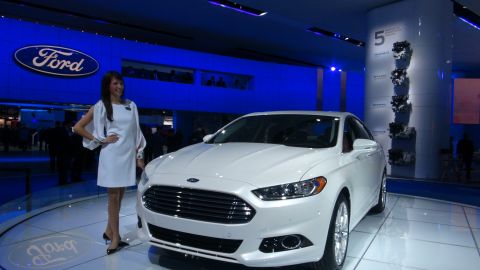 1993 Ford Taurus - A woman stands next to a white Ford sedan at an auto show with the Ford logo displayed prominently.