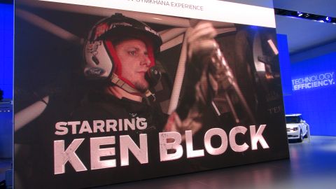 1993 Ford Taurus - A white car is displayed at an event with a large screen showing a video of Ken Block.