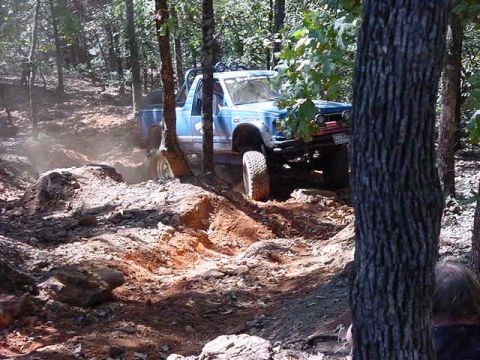 1986 Ford Bronco - A blue off-road vehicle tackles a challenging dirt path surrounded by trees.