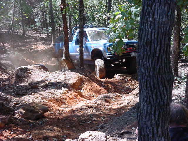 Lifted light blue pickup with roof light bar climbing a rocky red-dirt trail obstacle in the woods