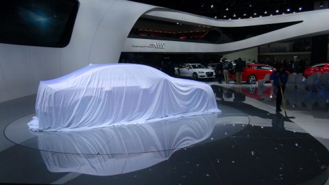 1993 Ford Taurus - A car covered by a white sheet is displayed on a platform at an auto show, with other vehicles and attendees in the background.