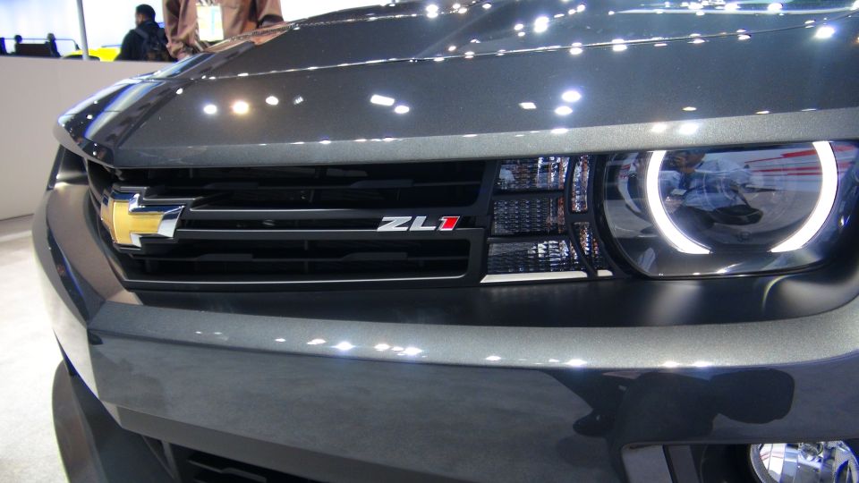 Gray Chevrolet Camaro ZL1 front grille with a gold bowtie emblem and ZL1 badge, next to a headlight with a white halo ring