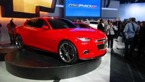 1993 Ford Taurus - A red concept car is showcased at an auto show, with people observing and a large digital display in the background.