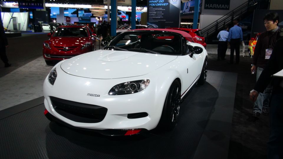 White Mazda convertible with red accents on the front bumper, parked indoors at a car show. A red car is visible behind it