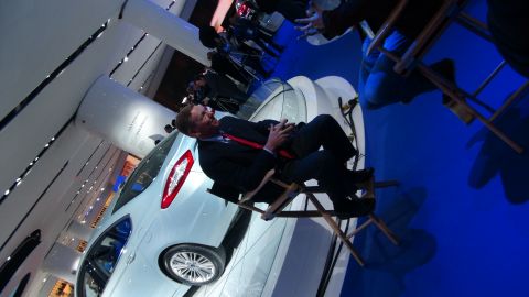 1993 Ford Taurus - A man in a suit is seated next to a white car at an auto show.