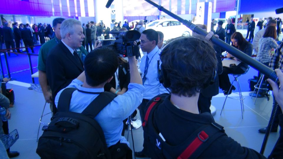 Media crew filming people at an auto show with a white car in the background