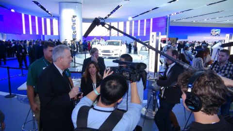 1993 Ford Taurus - A white Ford sedan is showcased at an auto show, with media personnel filming the event.