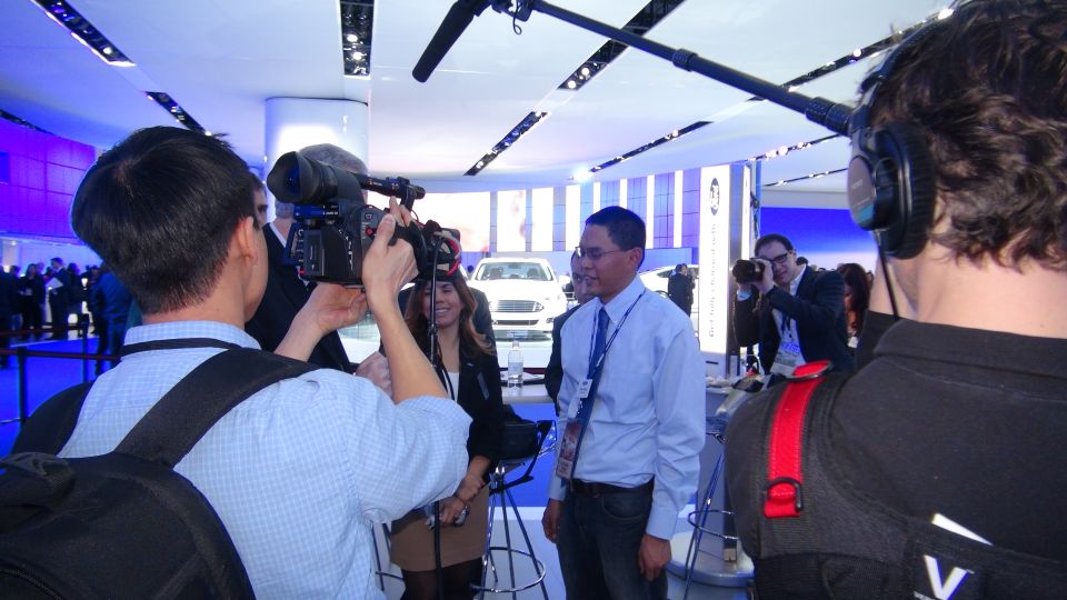 Media filming a man at an auto show with a white car in the background