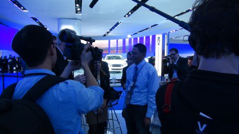1993 Ford Taurus - A white car is displayed at an auto show while media personnel film and photograph it.