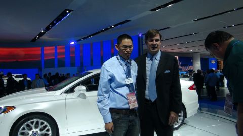1993 Ford Taurus - Two men stand next to a white Ford sedan at an auto show, with other vehicles and attendees in the background.