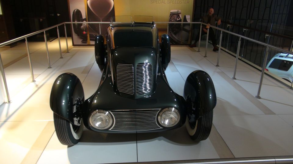 Front view of a dark green vintage speedster with exposed front wheels and a prominent chrome grille