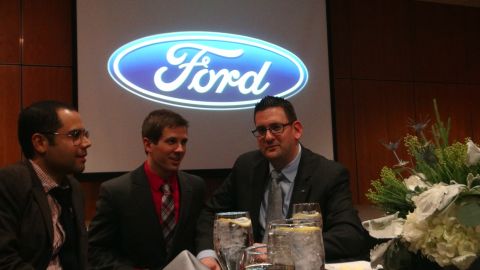 1993 Ford Taurus - Three men are seated at a table with drinks and a floral arrangement, with a large Ford logo projected on a screen behind them.