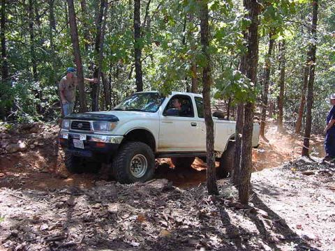 1986 Ford Bronco - A white pickup truck navigates a wooded off-road trail, kicking up dust.