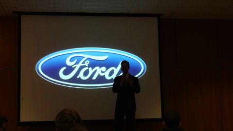 1993 Ford Taurus - A speaker stands in front of a projected Ford logo during a presentation.