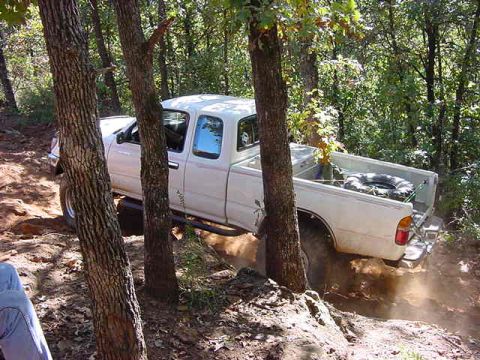 1986 Ford Bronco - A white pickup truck is seen off-roading on a dirt trail surrounded by trees, with dust being kicked up by its tires.