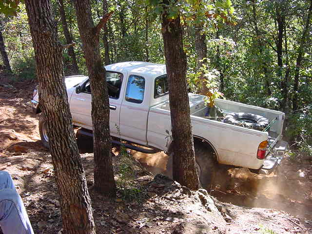 White Tacoma pickup with rock sliders climbing a dusty grade between trees