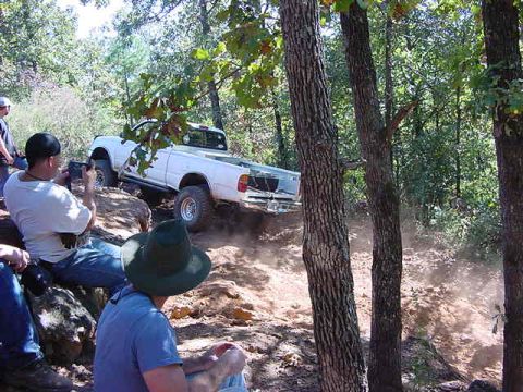 1986 Ford Bronco - Two white pickup trucks are seen off-roading on a dirt trail with spectators.