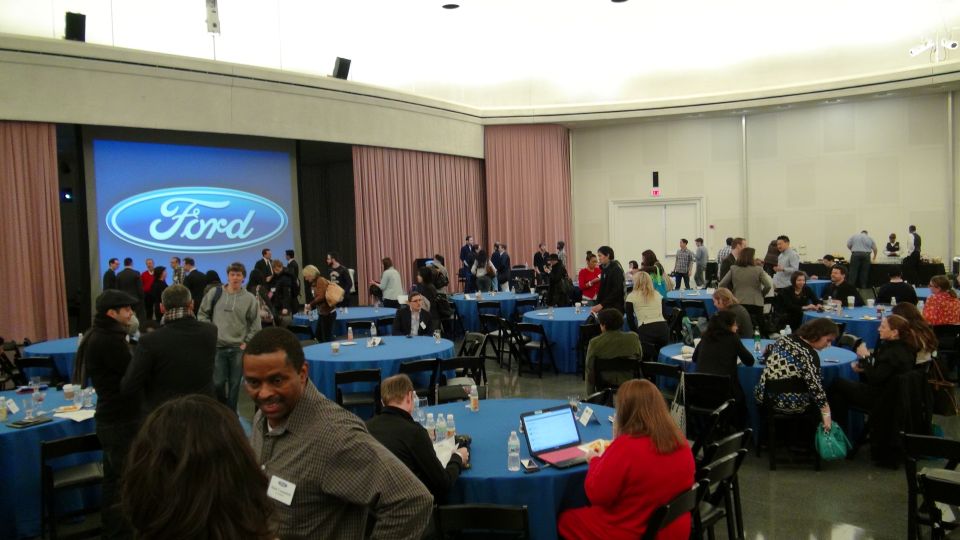 Ford logo displayed on a large screen in a conference room with people seated at blue-clothed tables