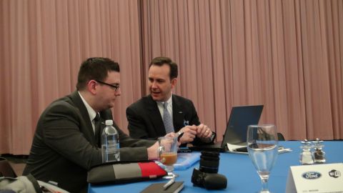 1993 Ford Taurus - Two men in suits are seated at a table, engaged in conversation, with a laptop and camera visible on the table.