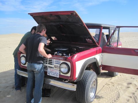 1968 Ford Bronco - Two men examine the engine of a red and white 1968 Ford Bronco parked on a sandy terrain under a clear blue sky.