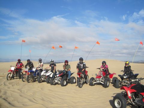 1968 Ford Bronco - A group of riders on various off-road vehicles pose in the sand dunes under a partly cloudy sky.