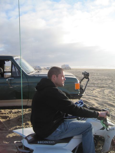 1968 Ford Bronco - A man rides an ATV on a sandy beach next to a green truck under a cloudy sky.